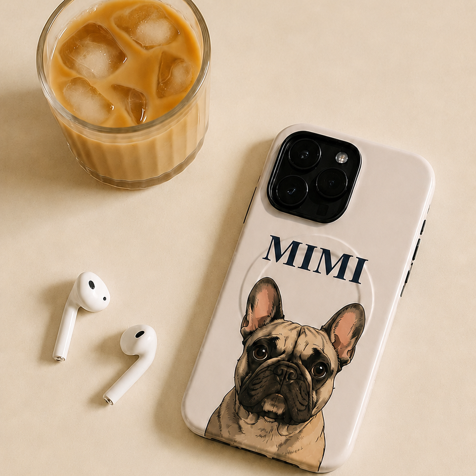A beige phone case featuring a printed illustration of a French Bulldog named 'MIMI' in black text, displayed on a beige surface next to an iced coffee, earbuds, and a smartphone.