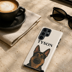 A white phone case featuring a custom German Shepherd illustration and the name 'TYSON' printed in black, displayed on a textured stone surface next to a cup of coffee and sunglasses.
